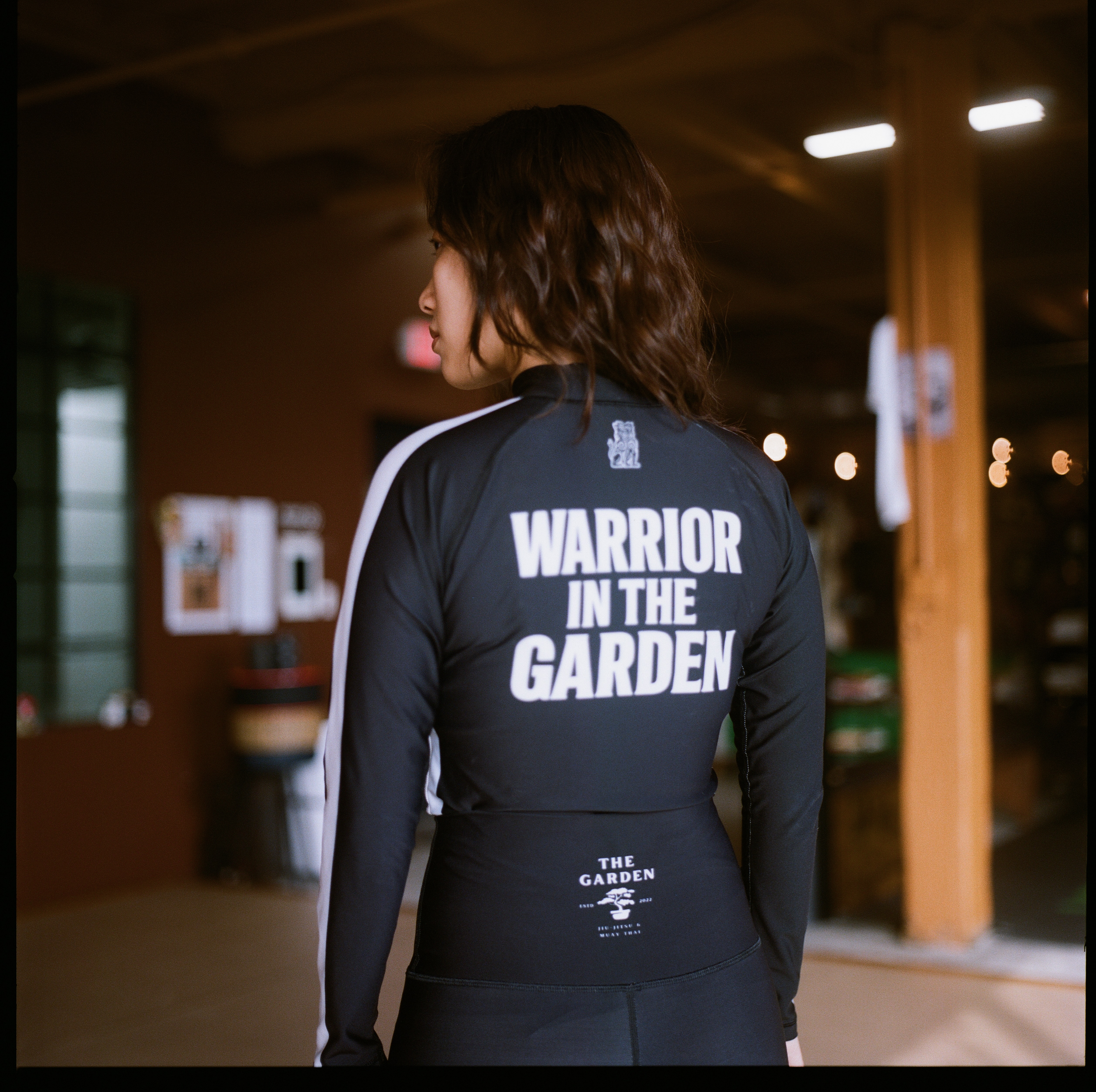 The new black Garden rashguard worn at the women’s no-gi camp, seen from behind with WARRIOR IN THE GARDEN across the shoulders and The Garden bonsai mark at the waist.