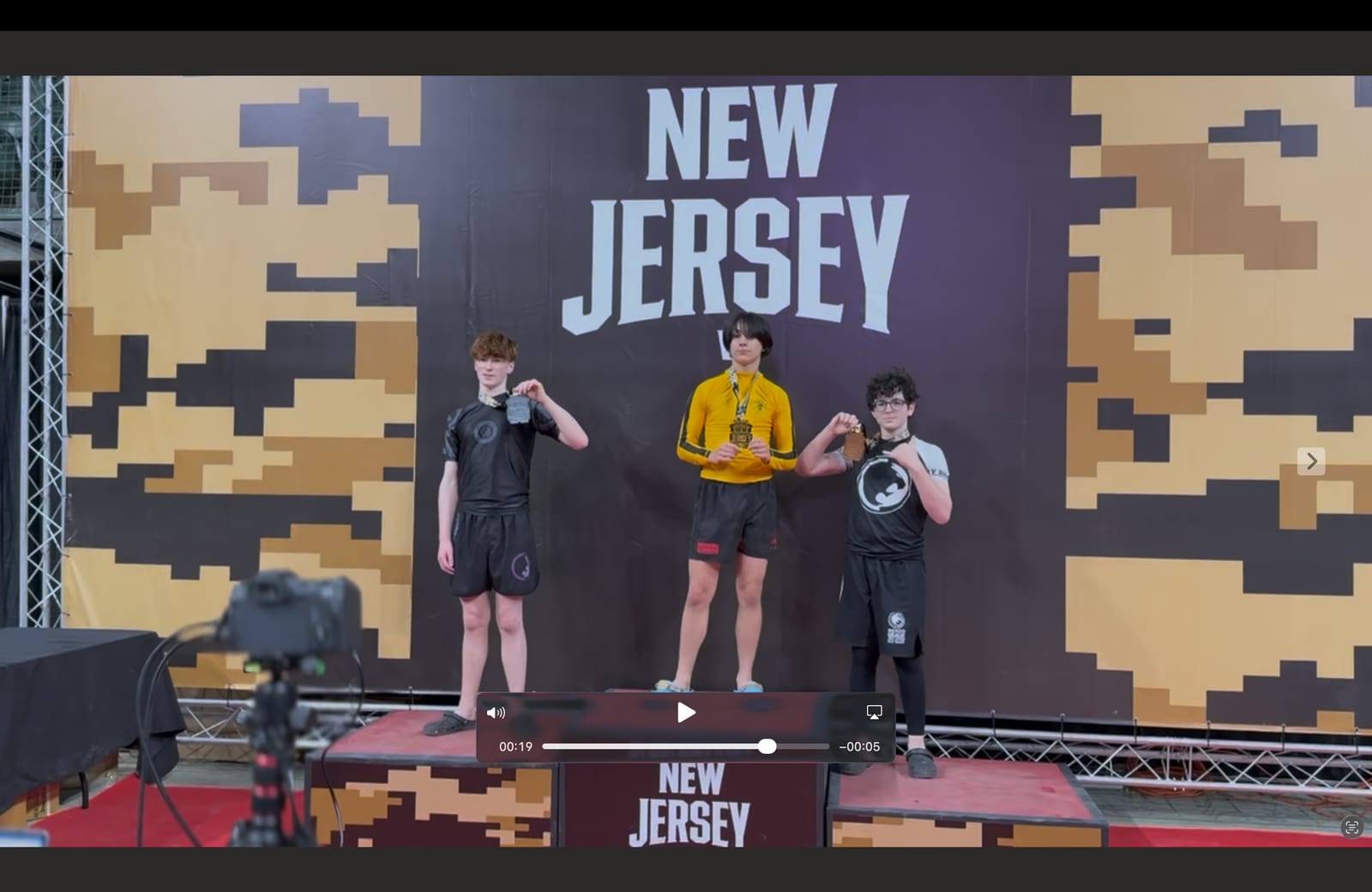 Three teenage grapplers on the medal podium at a New Jersey jiu-jitsu tournament, each holding up the medal hanging from their neck in front of a camo-patterned NEW JERSEY banner