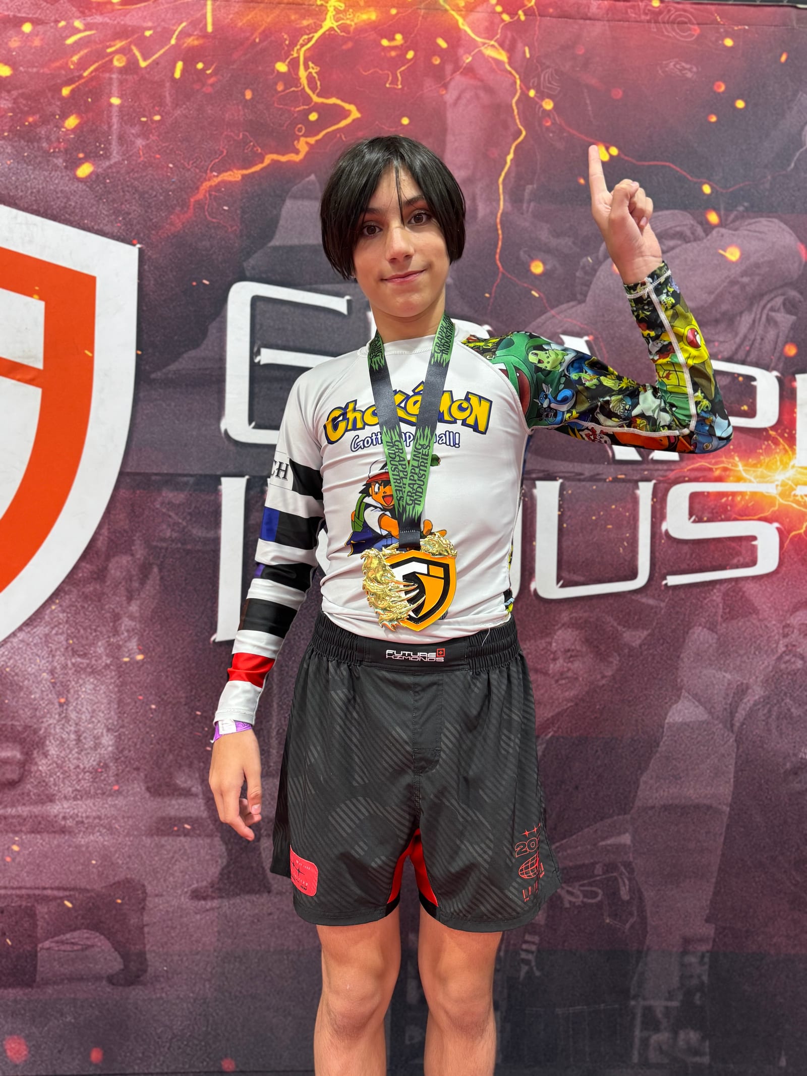 Three teenage grapplers on the medal podium at a New Jersey jiu-jitsu tournament, each holding up the medal hanging from their neck in front of a camo-patterned NEW JERSEY banner